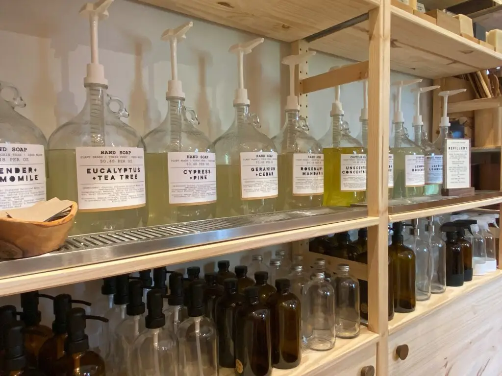 Refill station with large soap dispensers and empty glass bottles on wooden shelves.